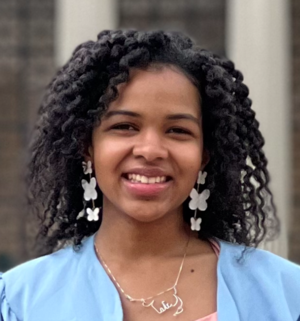 Tatianna is a Haitian-American woman with curly shoulder-length hair. She is smiling and wearing butterfly earrings in front of a blurred pillared background.